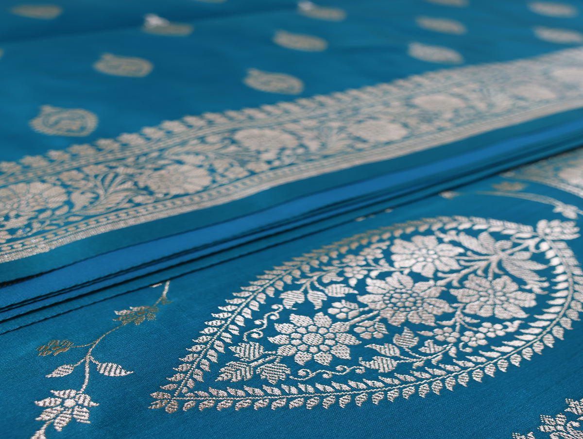 A close-up of a teal blue fabric, likely a Banarasi silk saree body, featuring intricate silver zari floral and paisley motifs woven across the surface.
